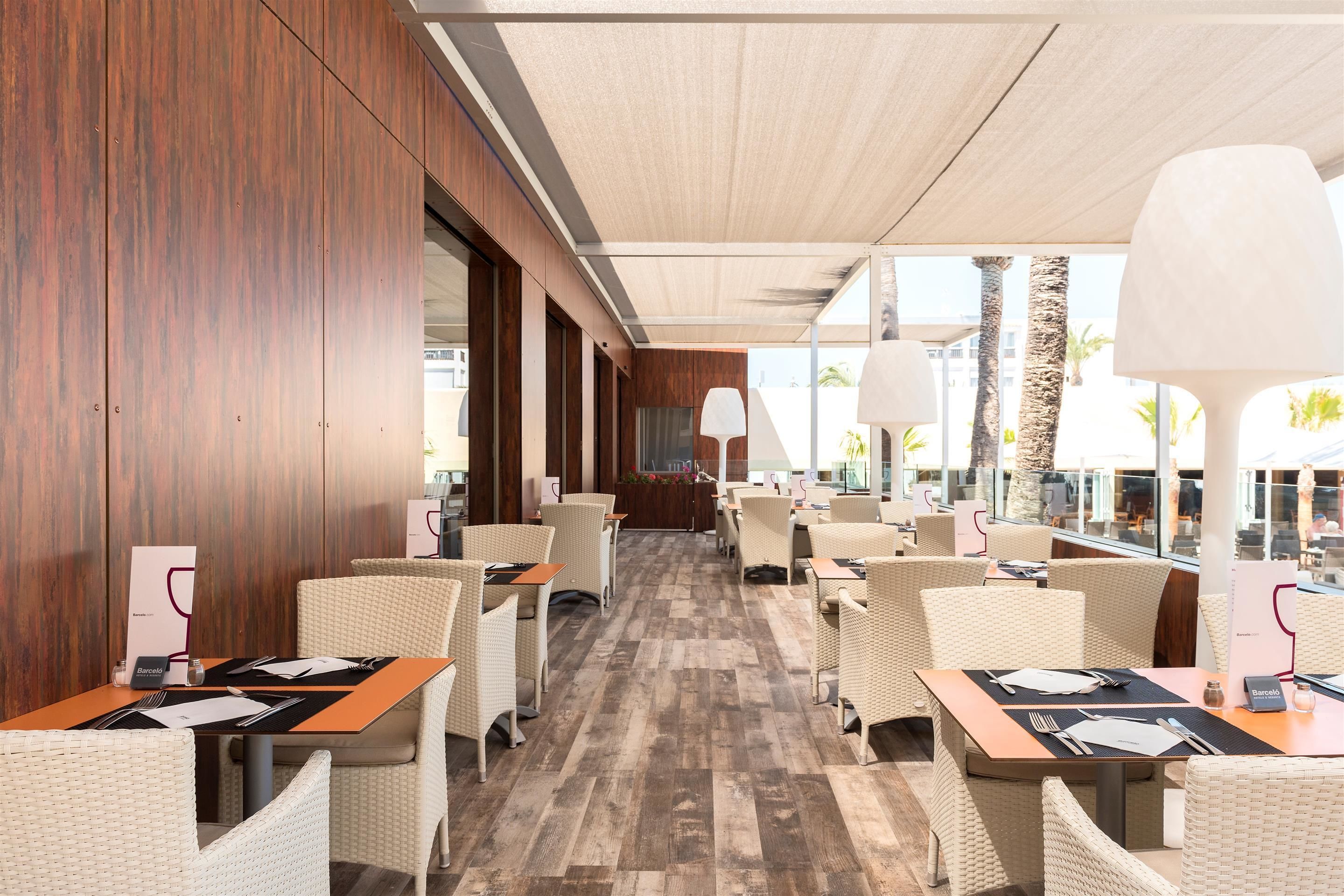 Een modern restaurant met rieten stoelen, houten vloeren en grote ramen met uitzicht op palmbomen en gebouwen.