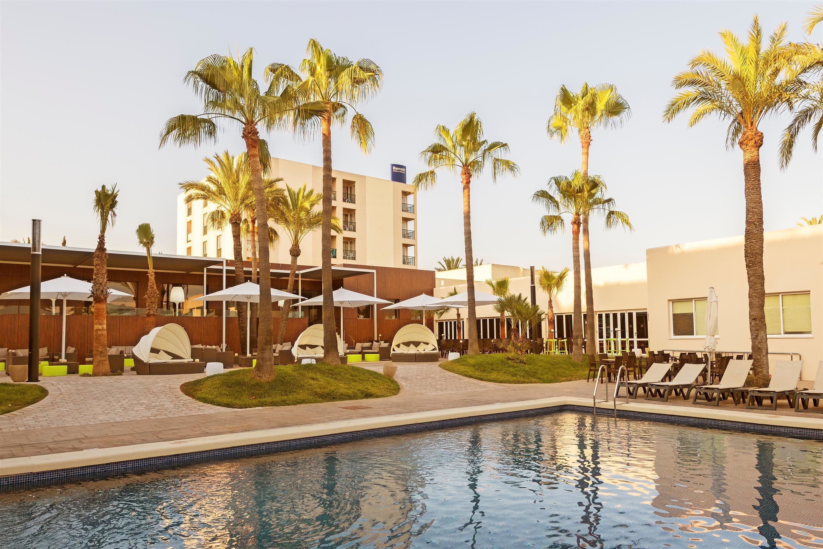 Zona de piscina de hotel con palmeras, sillones de descanso y sombrillas.