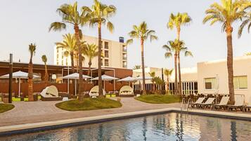 Zona de piscina de hotel con palmeras, sillones de descanso y sombrillas.