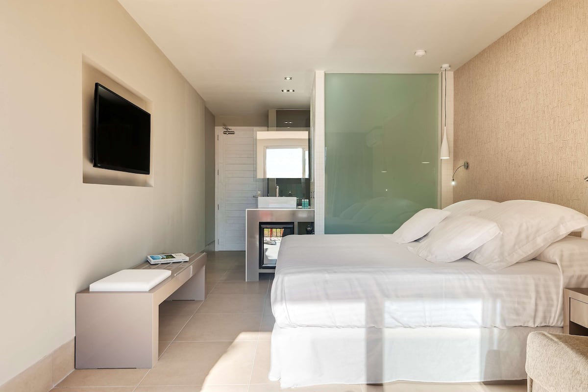 Une chambre moderne avec un lit, une table de chevet, un téléviseur fixé au mur et une salle de bain visible à travers une porte en verre.