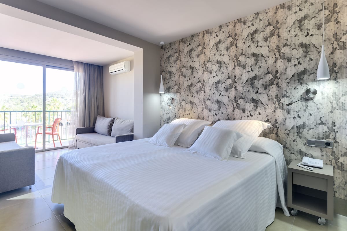 Une chambre d’hôtel moderne avec un grand lit, un balcon avec une chaise et une table de chevet avec une lampe.
