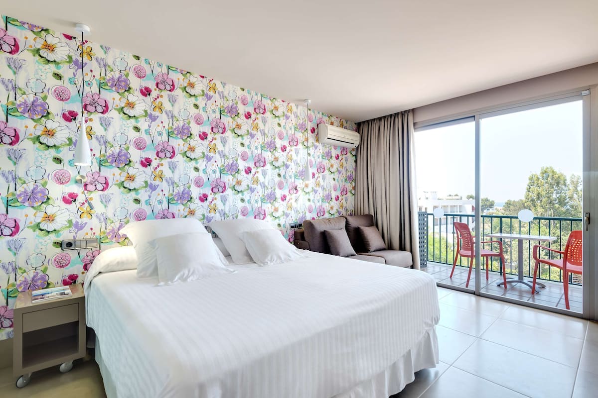 Une chambre d’hôtel avec un grand lit, une table de chevet, une chaise, un balcon avec des chaises rouges et un papier peint à motifs floraux.