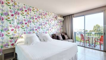 Quarto de hotel com uma cama grande, uma mesinha de cabeceira, uma cadeira e uma varanda com cadeiras vermelhas, além de papel de parede floral.