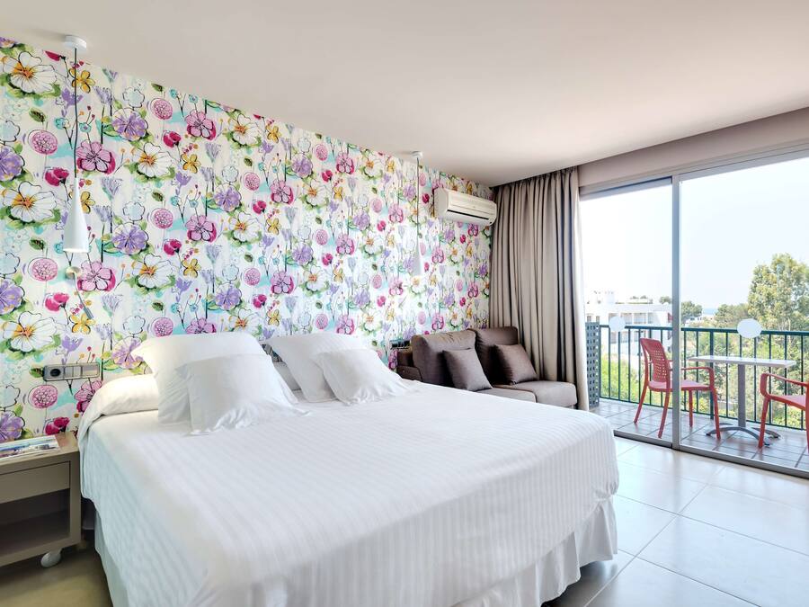 Quarto de hotel com uma cama grande, uma mesinha de cabeceira, uma cadeira e uma varanda com cadeiras vermelhas, além de papel de parede floral.
