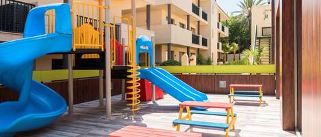 Zona de juegos infantiles con toboganes y mesas, rodeada por un edificio de varios pisos con balcones.