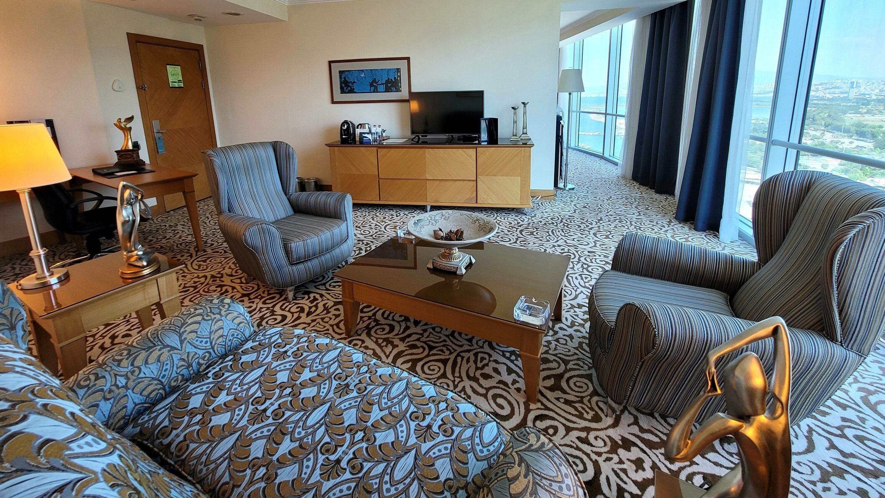 Habitación de hotel con alfombra estampada, un sofá, dos sillones, una mesa de centro, un televisor y un armario.