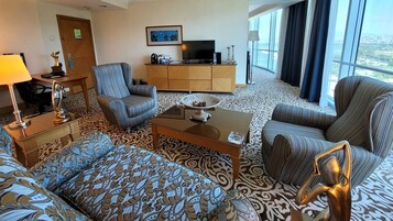 Habitación de hotel con alfombra estampada, un sofá, dos sillones, una mesa de centro, un televisor y un armario.