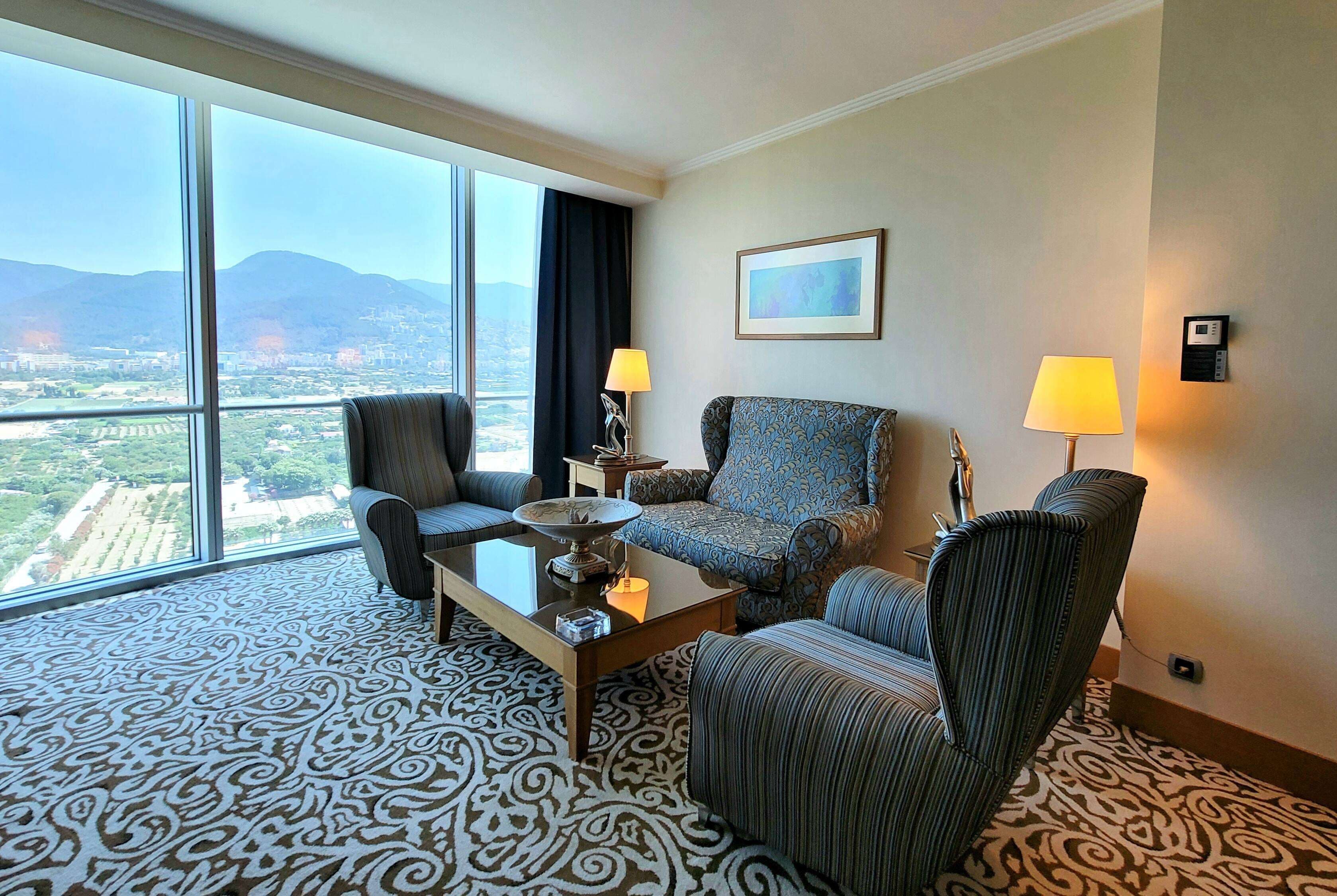 A hotel room with a large window, a patterned carpet, a sofa, two armchairs, a coffee table, and two lamps.