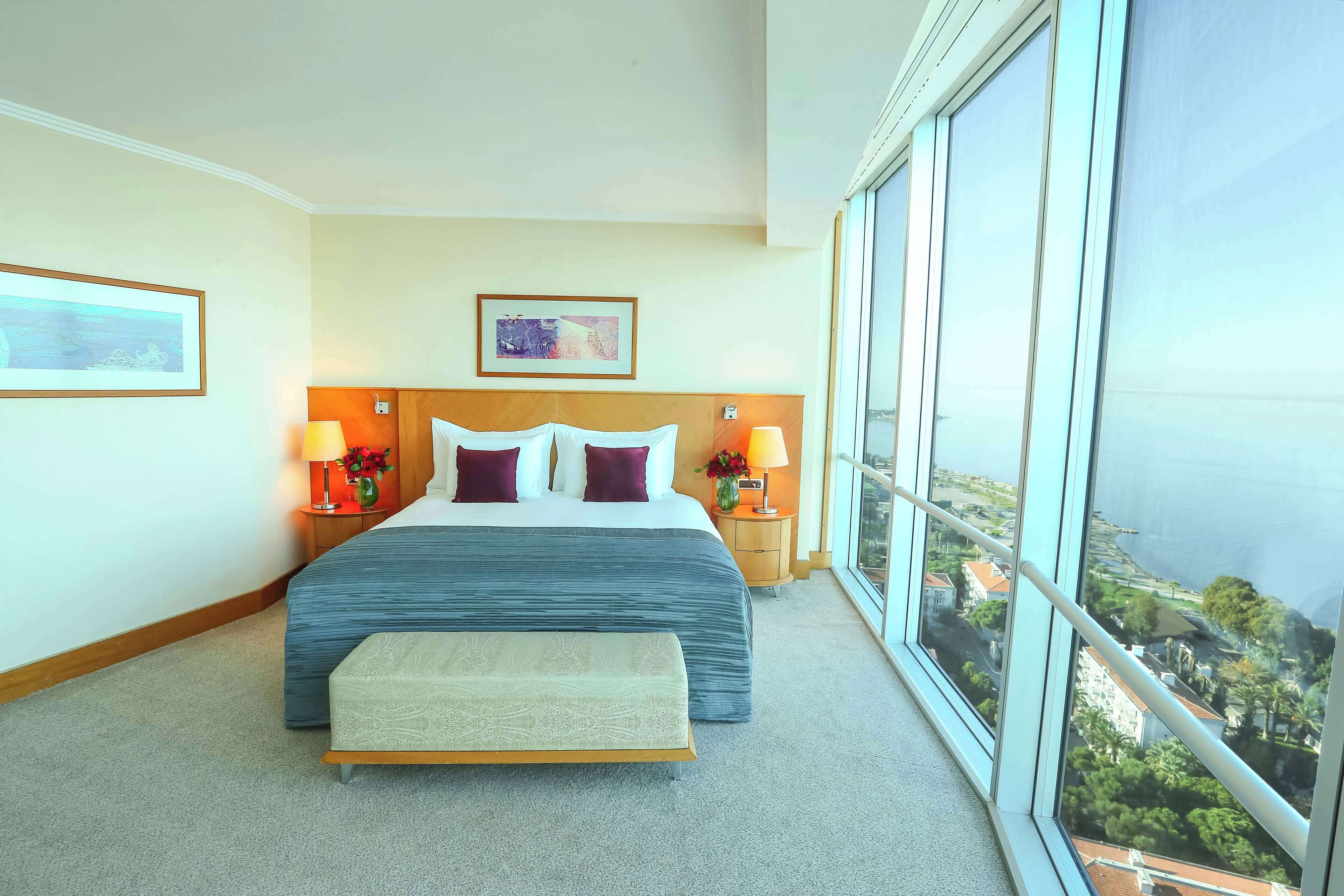 Habitación de hotel con una cama grande, dos mesitas de noche con lámparas, un cabecero y un ventanal con vistas a la ciudad.