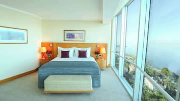 Quarto de hotel com uma cama grande, duas mesas de cabeceira com luminárias, cabeceira e uma janela ampla com vista para a cidade.