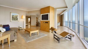 Een ruime woonkamer met een bank, een salontafel en een fauteuil. Een flatscreen-televisie is aan de muur bevestigd, en er is een groot raam met uitzicht op zee.