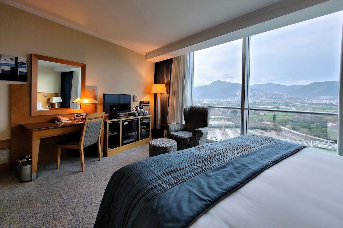 A hotel room with a large bed, a desk with a computer, a chair, a TV, and a view of a city and mountains.