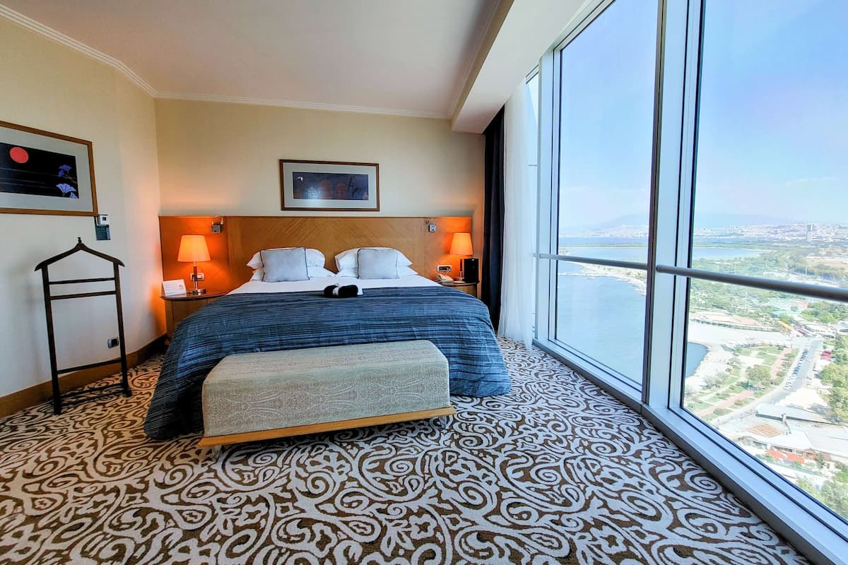 A hotel room with a large bed, two bedside lamps, a headboard, a hanging coat rack, a bench, and a large window with a city view.