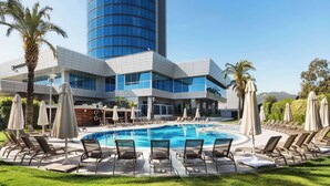 Een hotel met een zwembad, omringd door ligbedden en parasols, een modern gebouw met een glazen gevel en palmbomen.