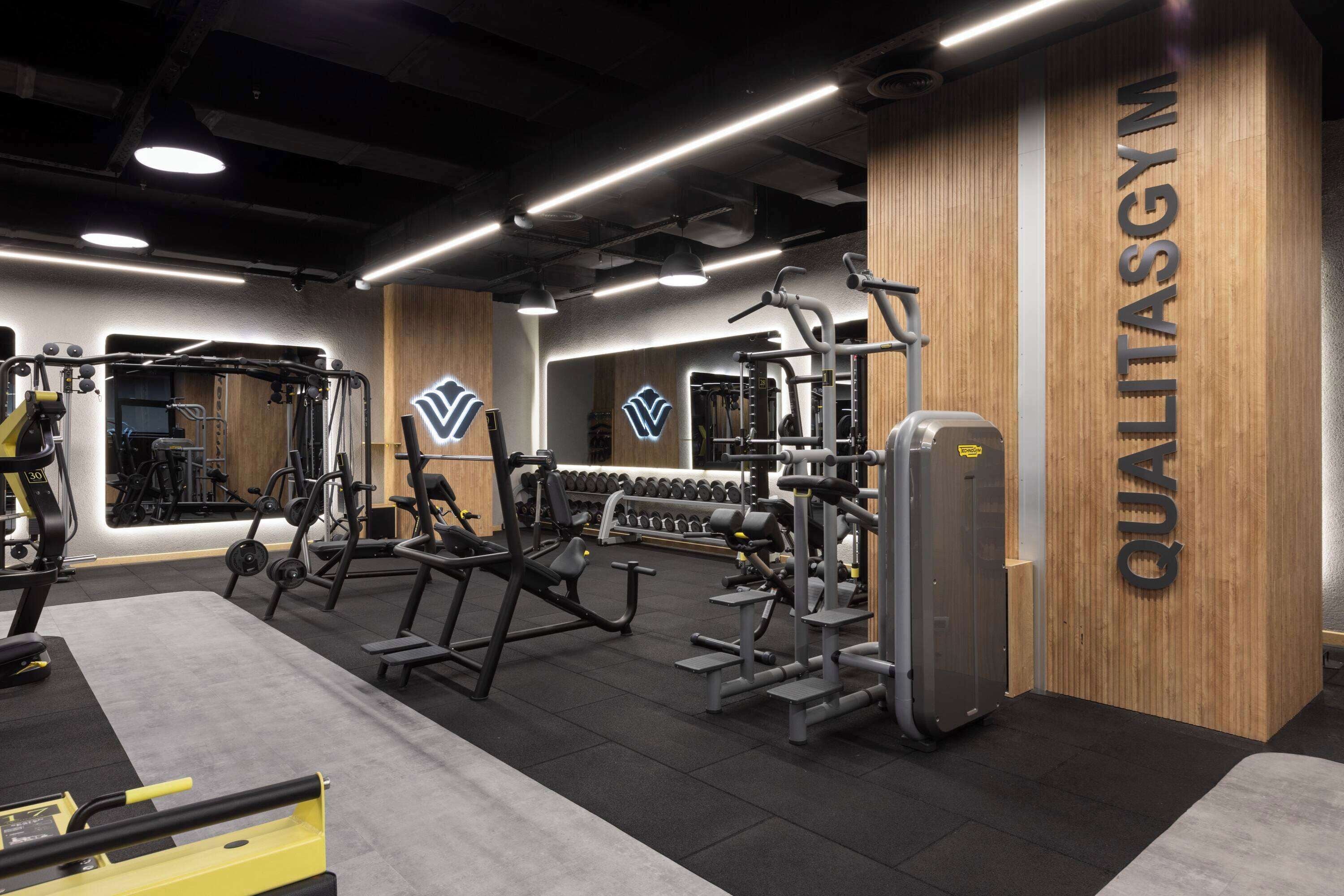 Un gimnasio con diversos equipos de ejercicio, un espejo grande y una pared de madera con el nombre 'QUALITASGYM'.