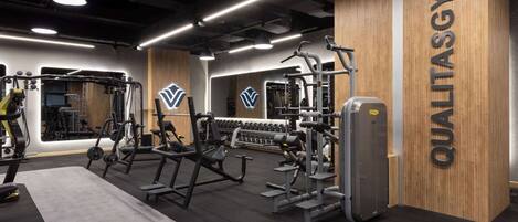 Un gimnasio con diversos equipos de ejercicio, un espejo grande y una pared de madera con el nombre 'QUALITASGYM'.
