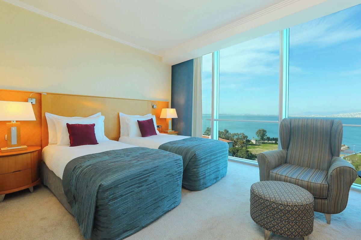 A hotel room with two beds, a large window with a view of the sea, a chair, and a small ottoman.