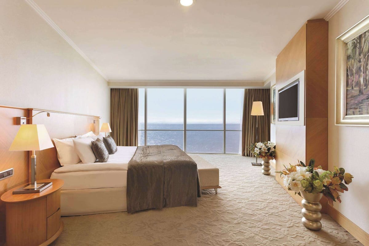 A hotel room with a large window offering a view of the sea, a bed with a headboard, bedside lamps, a bedside table, and a vase with flowers.