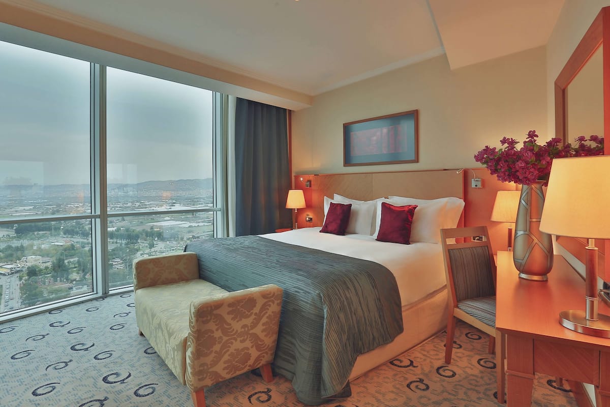 A hotel room with a large window, a bed, a chair, a desk, and a vase of flowers.