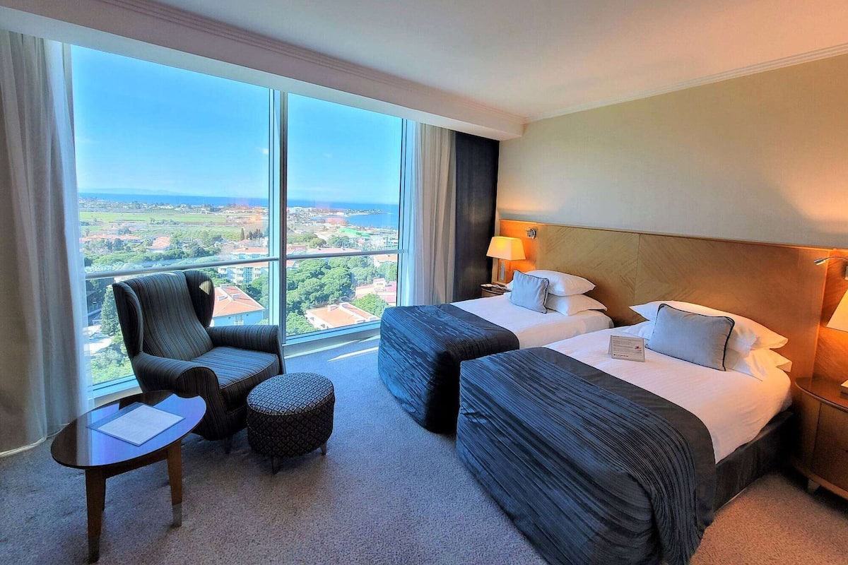 A hotel room with two beds, a chair, a small table, and a view of the city.
