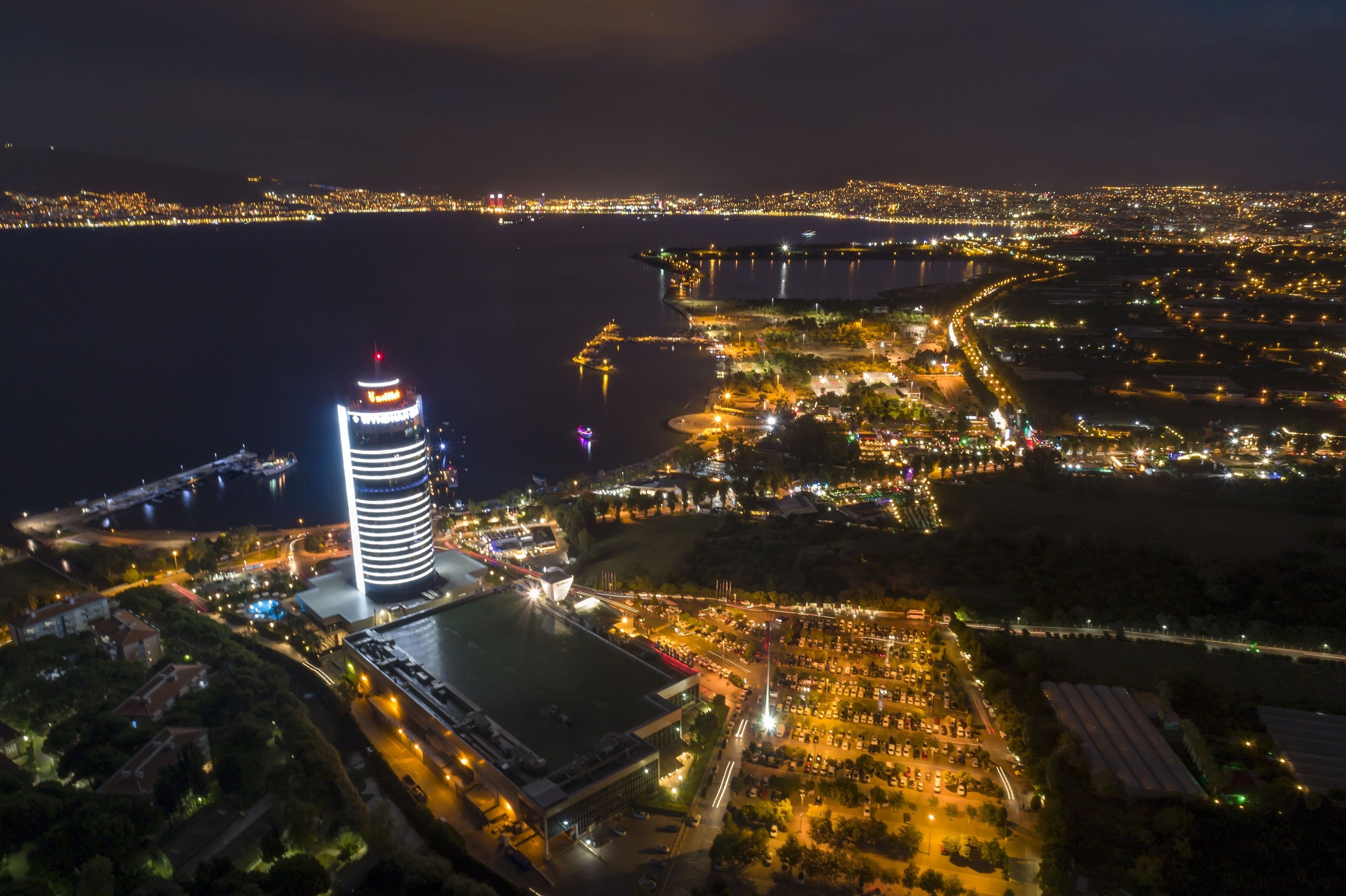 A nighttime aerial view of a coastal city with a prominent circular building, a marina, and a well-lit urban area.