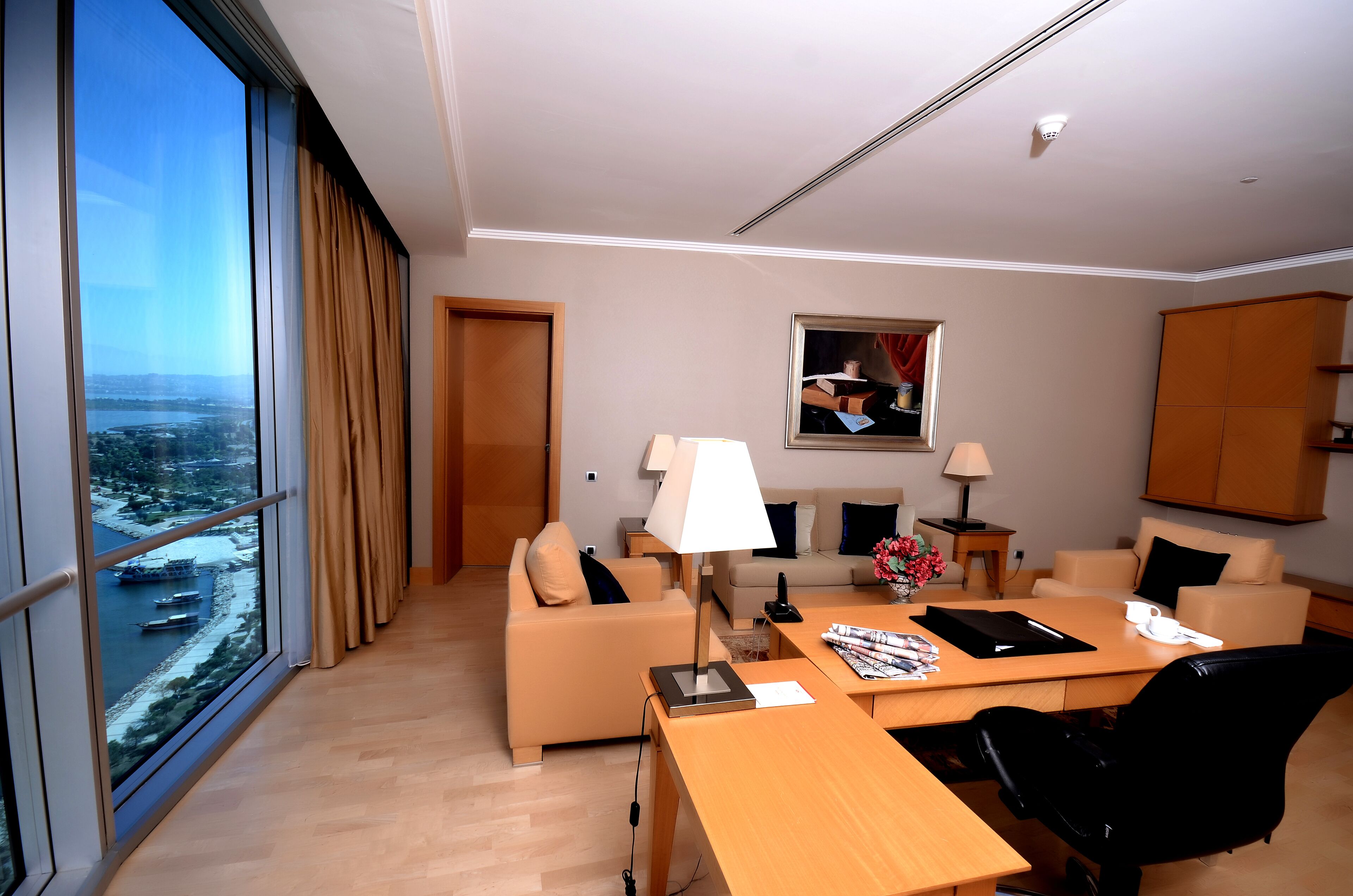 Habitación de hotel moderna con un gran ventanal que ofrece vista a la ciudad, un sofá, un escritorio con lámpara y una pequeña mesa con un florero.