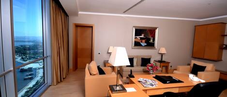 Habitación de hotel moderna con un gran ventanal que ofrece vista a la ciudad, un sofá, un escritorio con lámpara y una pequeña mesa con un florero.