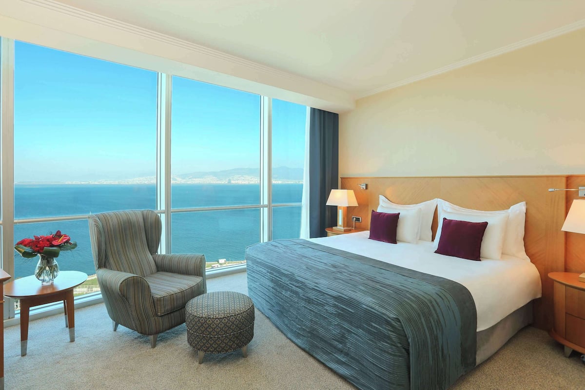 A hotel room with a large bed, a chair, a small table, and a view of the sea.