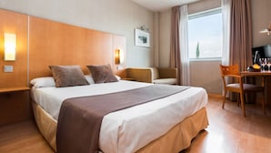 A hotel room with a large bed, a desk with a chair, a window with curtains, and a wall-mounted TV.