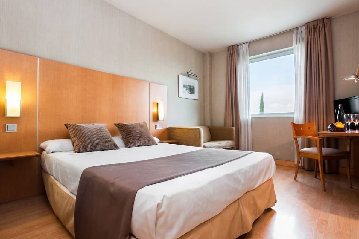 Ein Hotelzimmer mit einem großen Bett, einem Schreibtisch mit Stuhl, einem Fenster mit Vorhängen und einem an der Wand befestigten Fernseher.