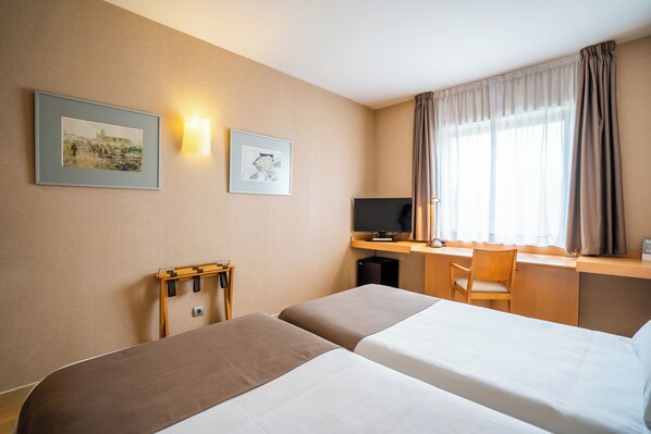 A hotel room with two beds, a desk with a computer, a chair, and two framed pictures on the wall.