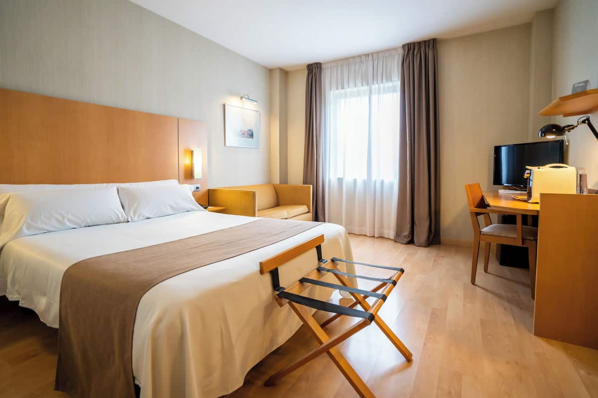 Ein Hotelzimmer mit einem großen Bett, einem Schreibtisch, einem Sessel, einem Fernseher und einem Fenster mit Vorhängen.