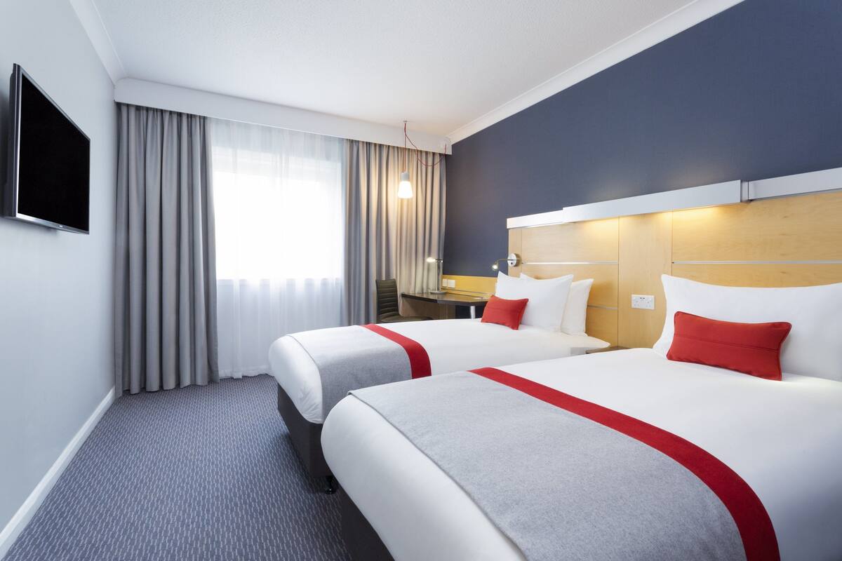 A hotel room with two beds, a TV, a desk, and a window with curtains.