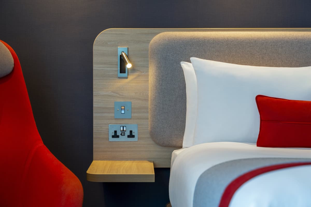 A modern hotel room with a wooden headboard, a bedside table, and a red chair.