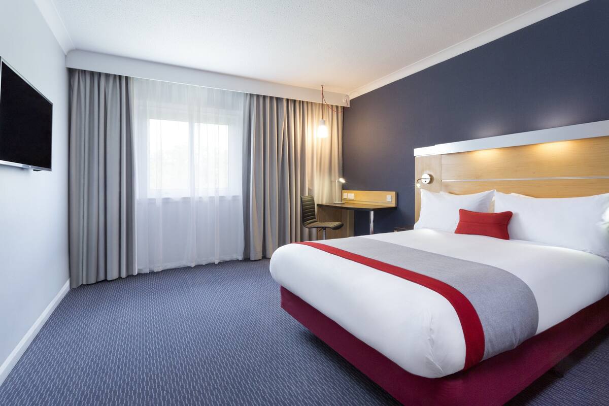 A hotel room with a large bed, a desk, a chair, a TV, and a window with curtains.