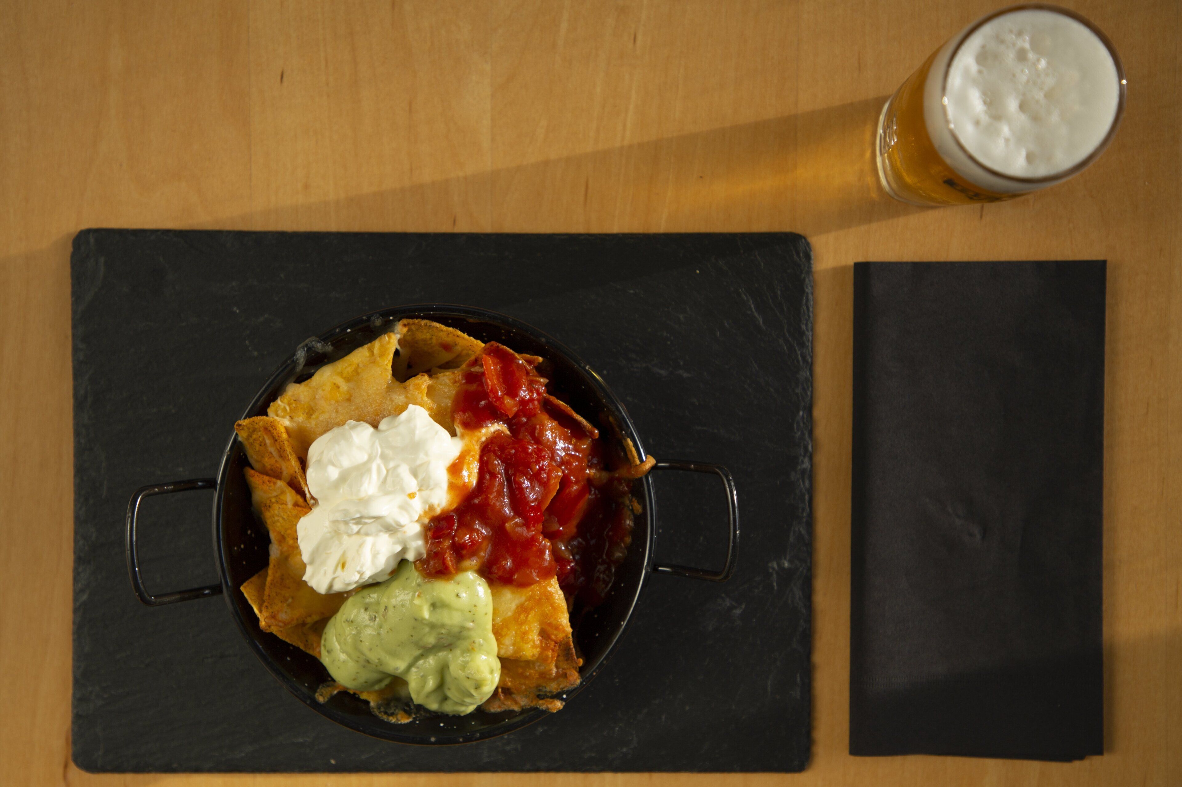 A meal consisting of nachos topped with sour cream, guacamole, and salsa, accompanied by a glass of beer on a slate coaster.