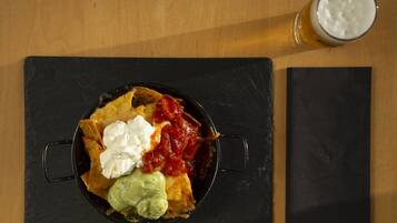 A meal consisting of nachos topped with sour cream, guacamole, and salsa, accompanied by a glass of beer on a slate coaster.