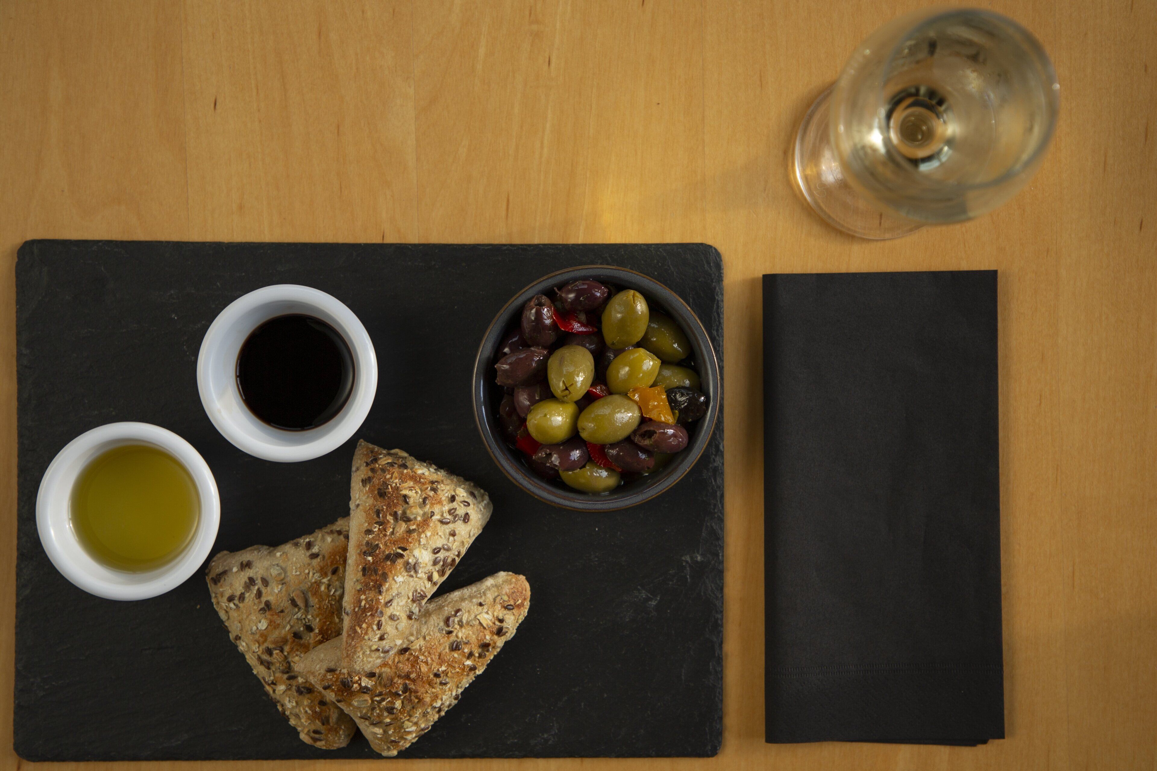 Una tabla de pizarra negra con un cuenco de aceitunas, dos recipientes pequeños de aceite de oliva y un trozo de pan sobre una mesa de madera.
