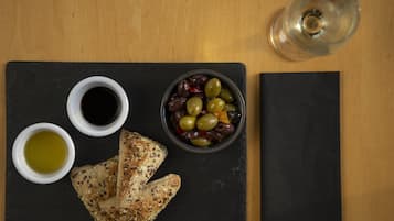 Una tabla de pizarra negra con un cuenco de aceitunas, dos recipientes pequeños de aceite de oliva y un trozo de pan sobre una mesa de madera.