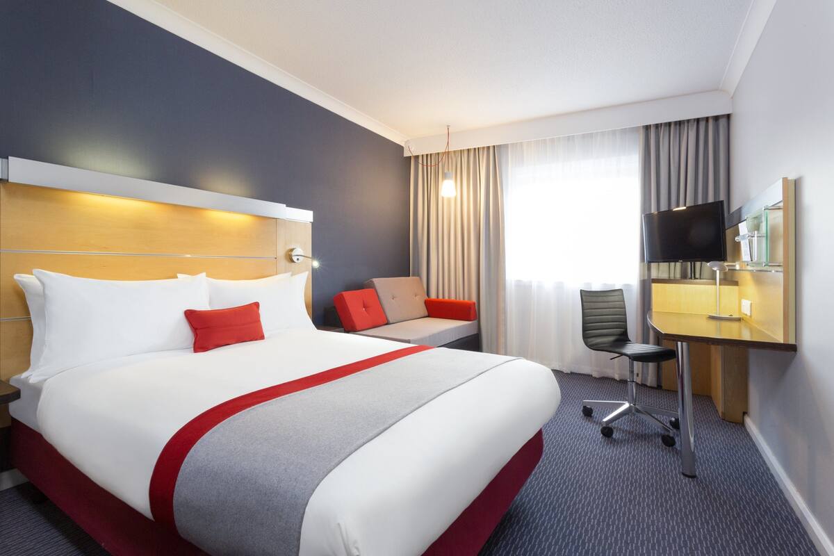 A hotel room with a large bed, a desk with a chair, a television, and a window with curtains.