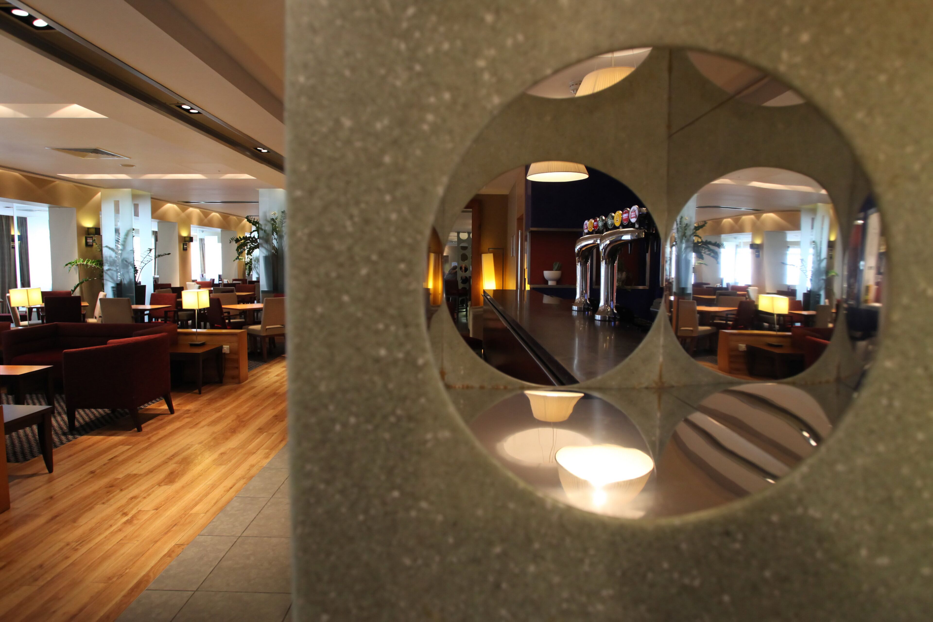 A modern hotel lobby with a circular mirror reflecting the interior, including a bar area and seating arrangements.