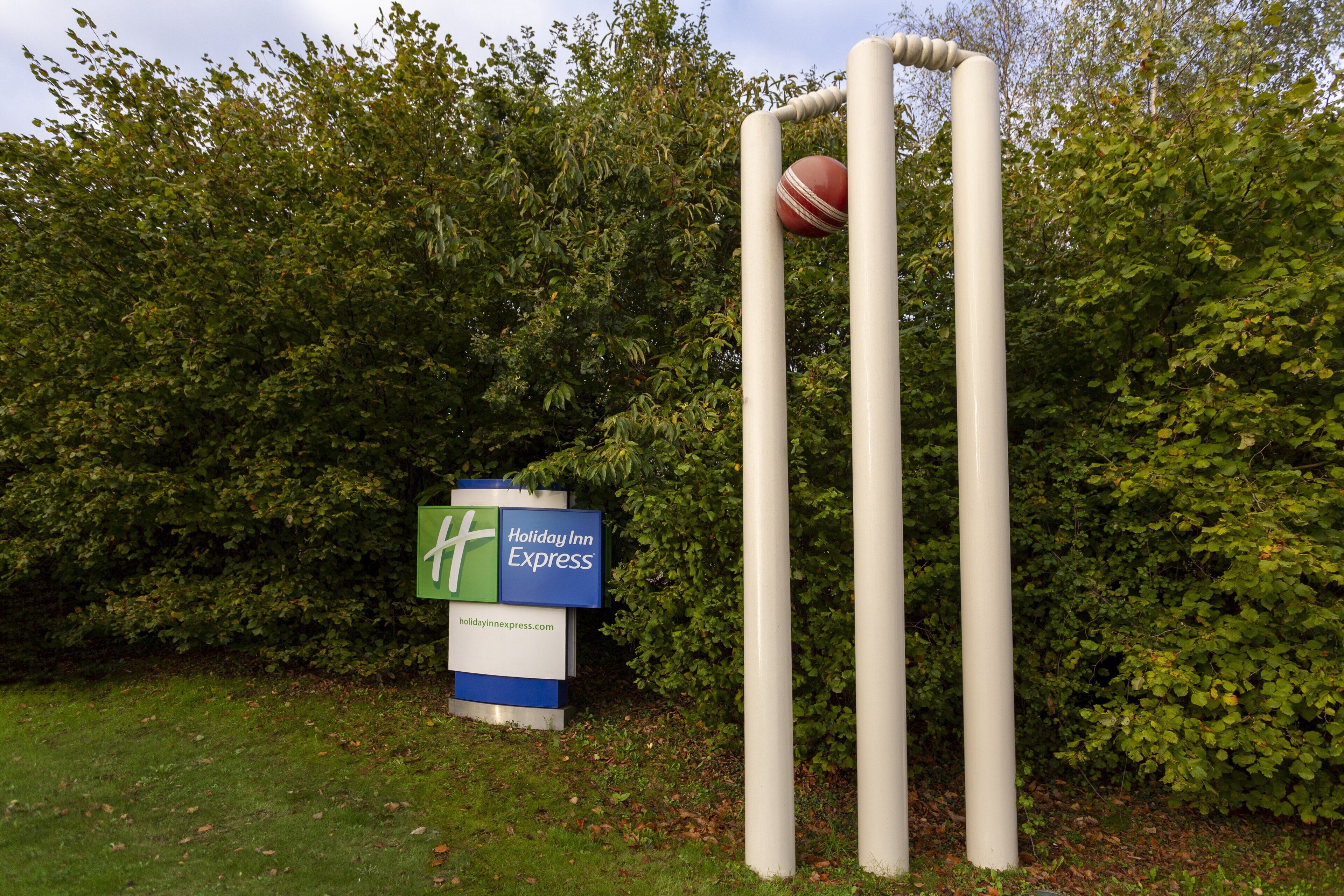 Un campo de cricket con tocones y pelota, y un cartel de Holiday Inn Express al fondo.