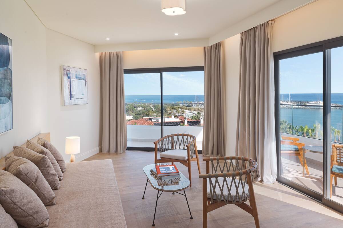 A spacious living room with a large window offering a view of the sea and a bridge. There is a sofa, two chairs, and a small table with a book.