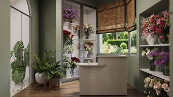 A room with a reception desk, shelves with flowers, and a large window with a view of greenery.