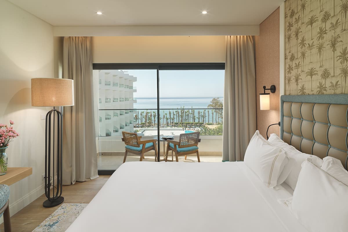 A hotel room with a large bed, a dining table with chairs, a view of the sea, and a balcony with a pool.