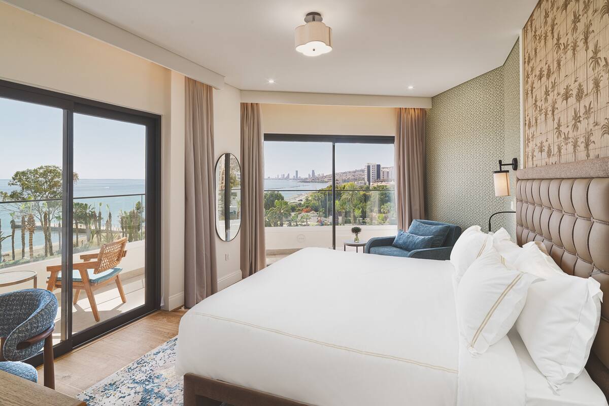 A hotel room with a large bed, a balcony with a view of the sea, and a cityscape in the background.