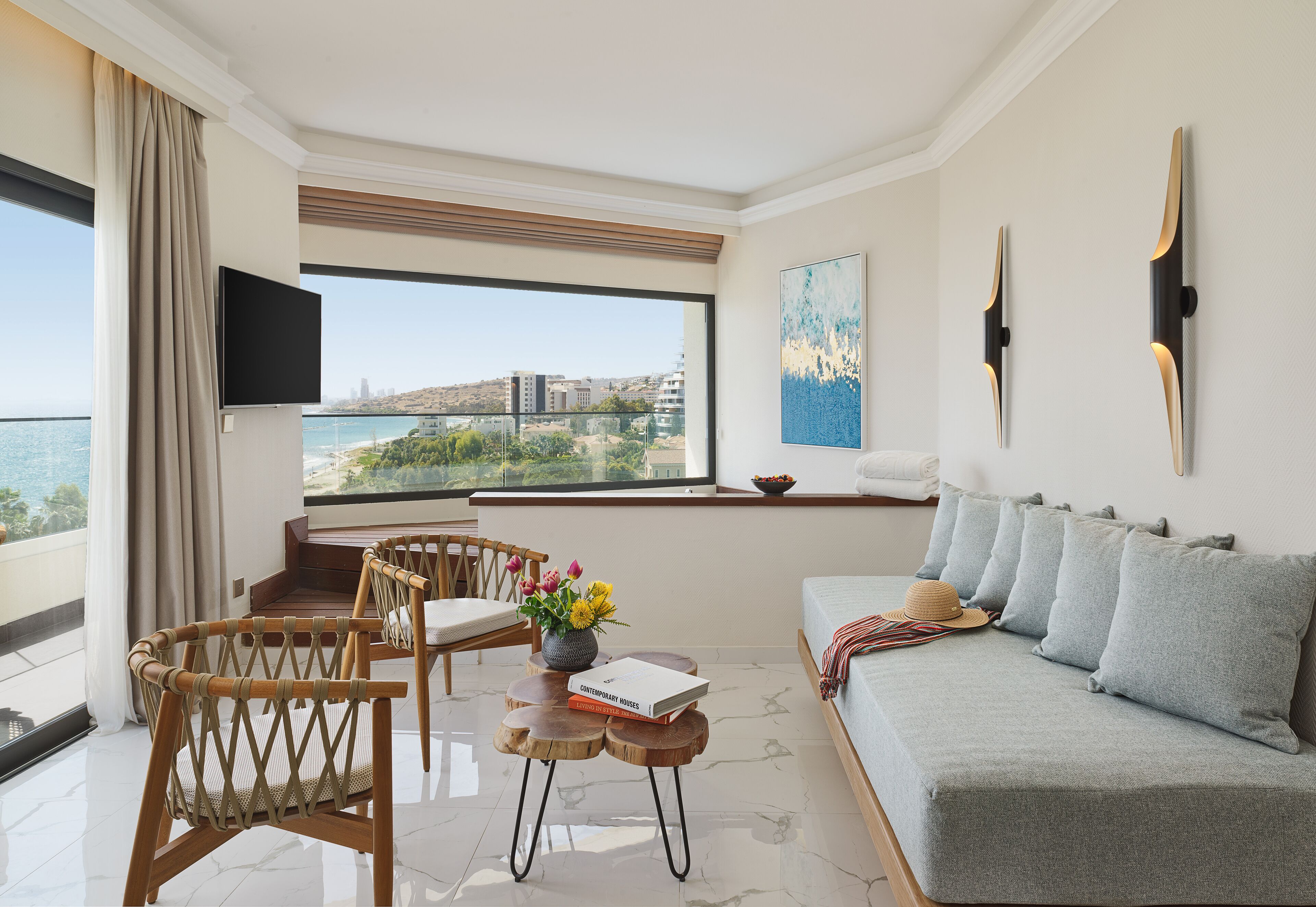 A modern living room with a sofa, chairs, a coffee table, and a TV. A large window offers a view of the city and sea.