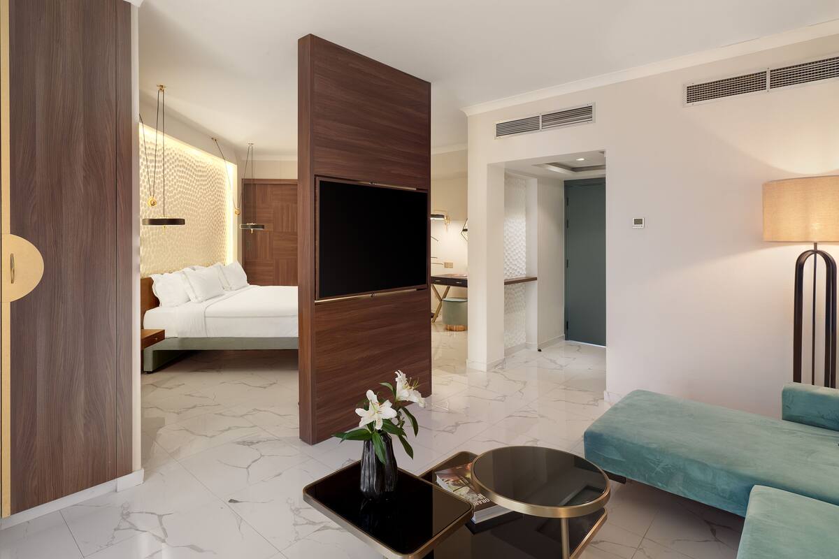 A modern hotel room with a bed, a blue sofa, a TV, and a small table with a vase of flowers.