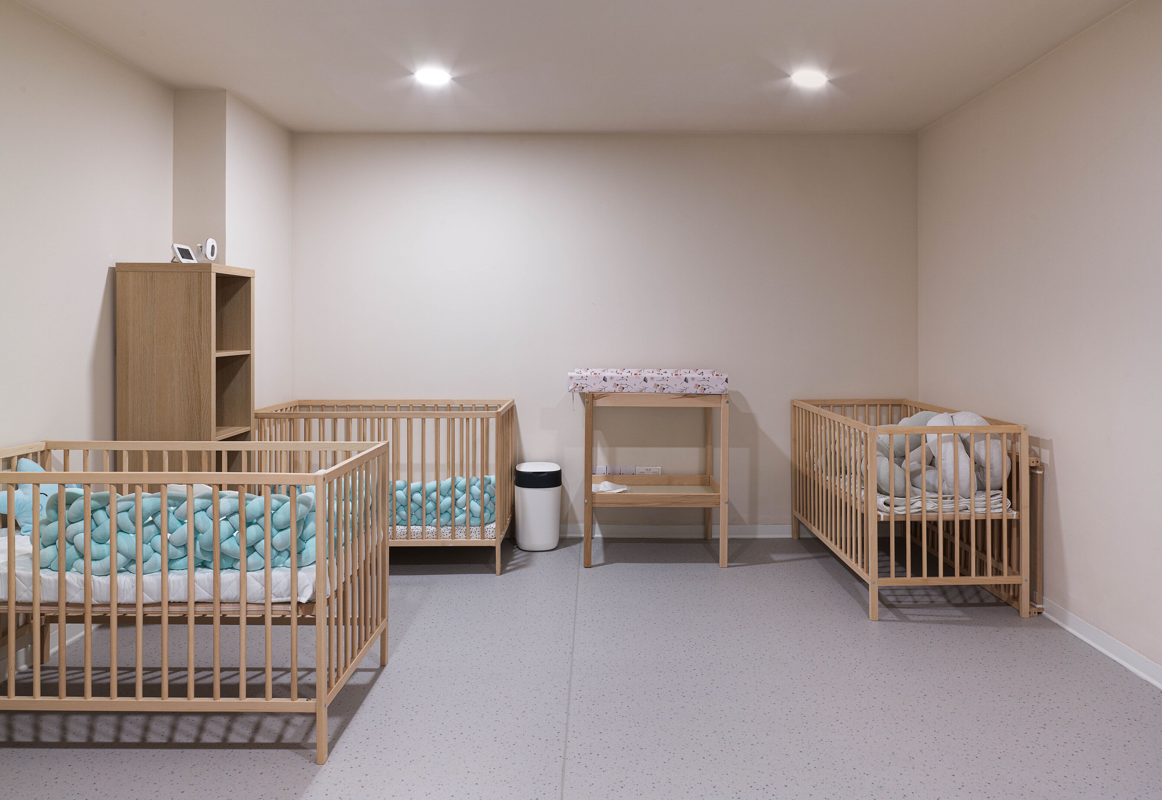 A room with two cribs, a wooden cabinet, and a small table.