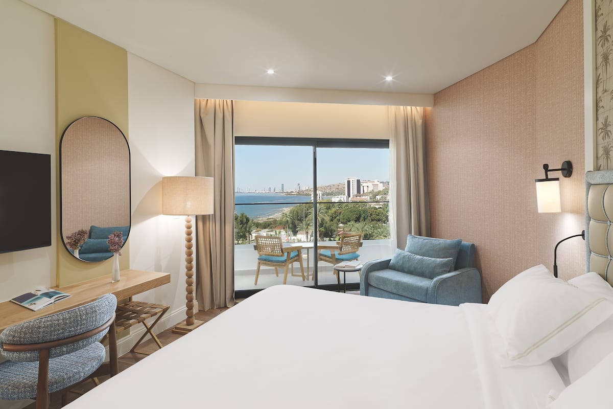 A hotel room with a large bed, a desk with a chair, a table with chairs, a view of the sea, and a cityscape.